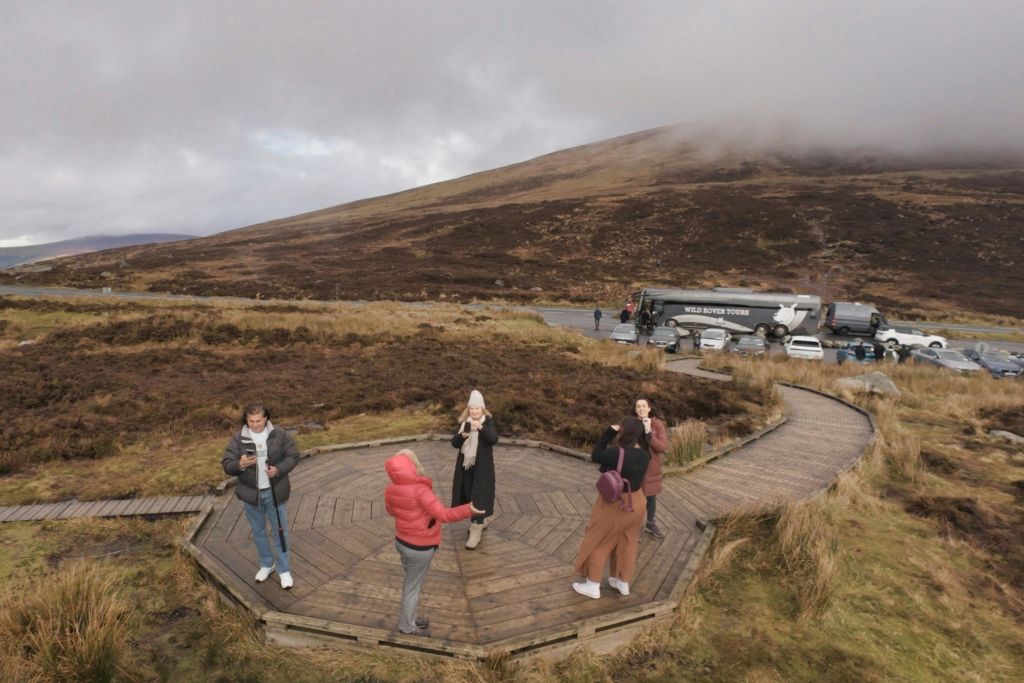 잔디가 우거지고 언덕이 많은 지역의 나무 플랫폼에 서 있는 한 무리의 사람들로, 근처에 투어 버스와 차가 주차되어 있습니다.
