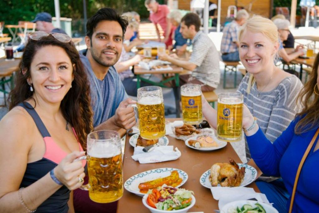 세 사람이 야외 테이블에 앉아 커다란 맥주잔을 들고 음식 접시를 앞에 두고 다른 사람들은 배경에 앉아 있습니다.