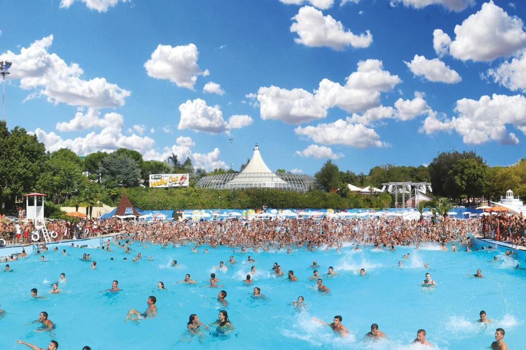 나무와 구조물로 둘러싸인 구름이 흩날리는 푸른 하늘 아래 사람들로 가득한 대형 야외 워터파크 수영장.