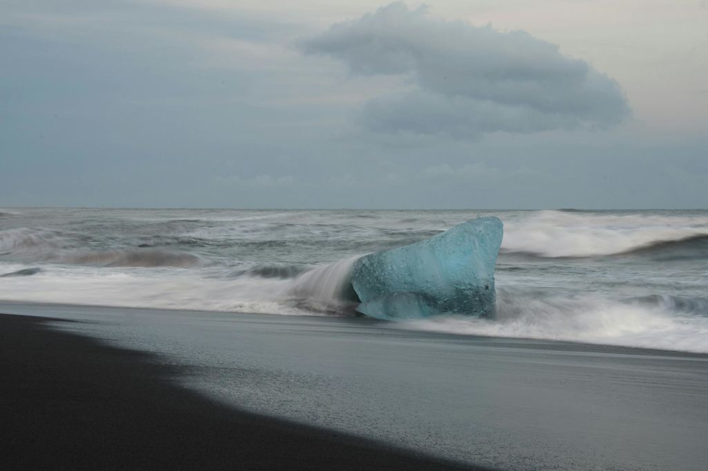 검은 모래 해변에 커다란 푸른 얼음 덩어리가 놓여 있고 흐린 하늘 아래 파도가 그 주위를 부딪칩니다.