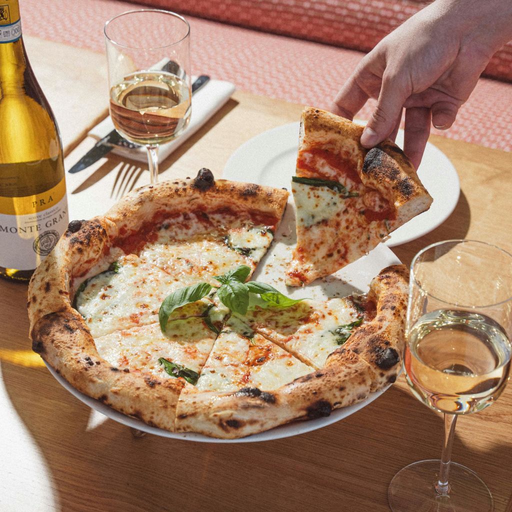 와인병, 화이트 와인 2잔, 식기가 놓인 나무 테이블에서 마르게리타 피자 조각을 손으로 들어 올립니다.