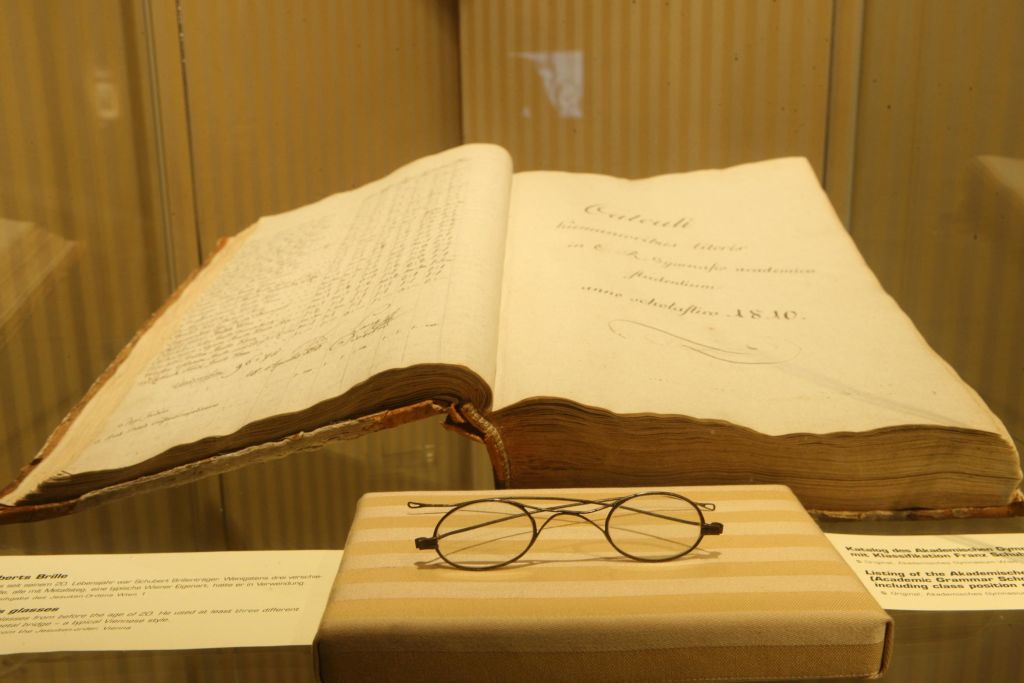 열려 있는 골동품 책이 유리 케이스에 전시되어 있으며, 그 아래 줄무늬 천에 둥근 와이어 테두리 안경이 놓여 있습니다.