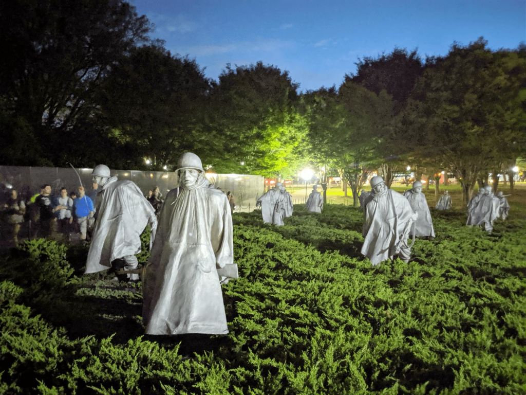 녹음이 우거진 가운데 우비를 입은 군인들의 조각상이 서 있으며, 해질 무렵 나무와 흐릿한 불빛이 배경을 이루고 있습니다.