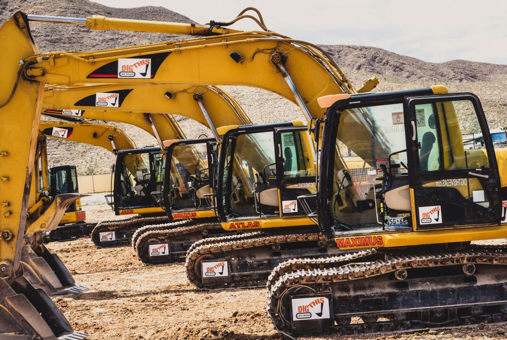 맑은 택시와 눈에 띄는 브랜딩이 있는 노란색 굴삭기가 줄지어 있으며, 산악 배경의 먼지에 주차되어 있습니다.