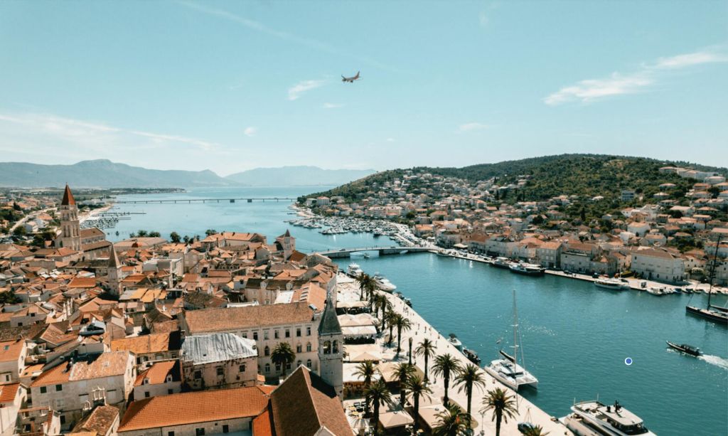 붉은 지붕의 건물, 야자수가 늘어선 바닷가, 정박한 보트, 머리 위로 날아가는 비행기가 있는 해안 마을입니다.