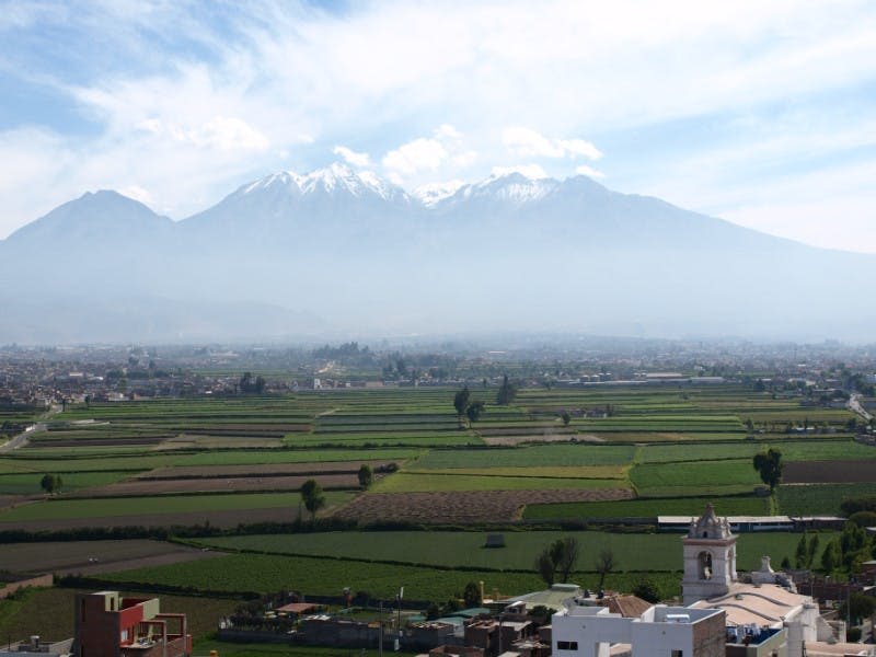 부분적으로 흐린 하늘 아래 푸른 들판과 흩어져 있는 건물, 산을 배경으로 한 풍경.