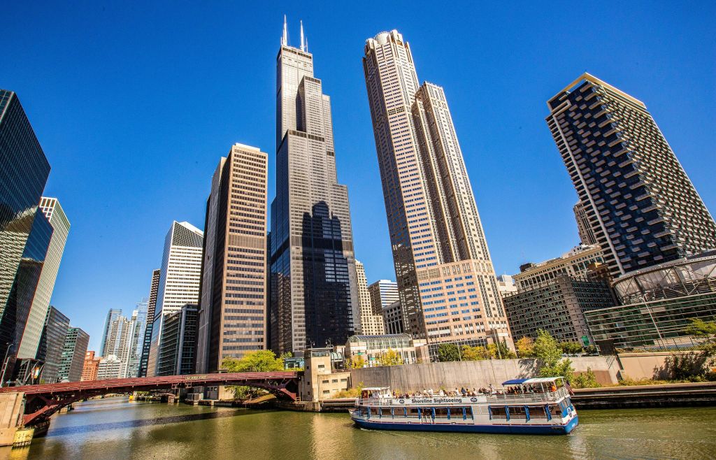 쌍둥이 안테나가 달린 키가 큰 검은색 안테나를 포함한 고층 건물은 맑고 푸른 하늘 아래 투어 보트가 있는 강을 따라 있습니다.