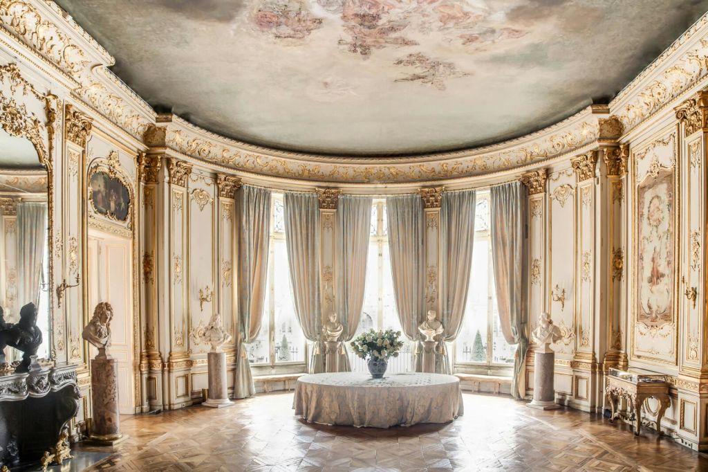 화려한 골드 디테일이 돋보이는 우아한 로툰다, 커튼으로 장식된 높은 창문, 조각상, 꽃무늬 센터피스가 있는 원형 테이블.