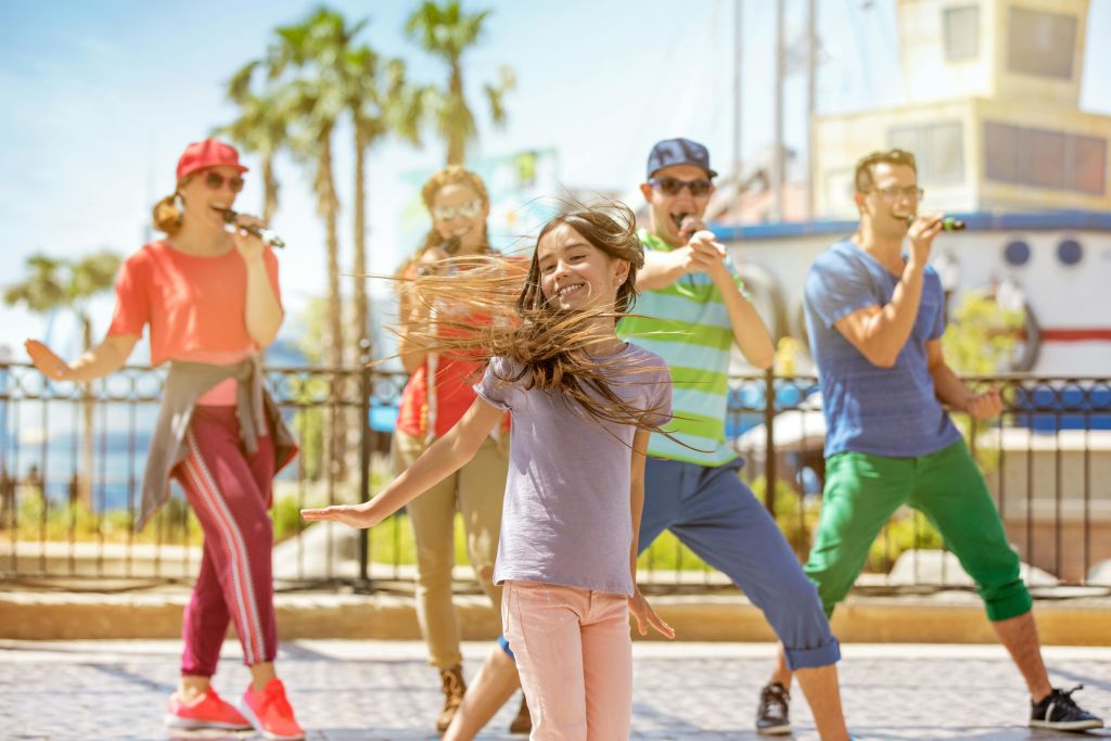 한 소녀가 머리카락을 휘날리며 즐겁게 춤을 추고, 뒤에서 네 명의 성인이 야자수가 있는 야외 환경에서 열정적으로 노래합니다.