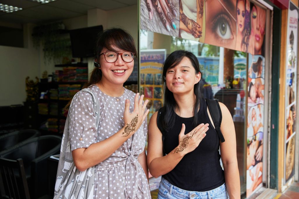 두 여성은 헤나 포스터와 함께 매장 밖에 서서 손에 복잡한 헤나 디자인을 자랑스럽게 보여줍니다.