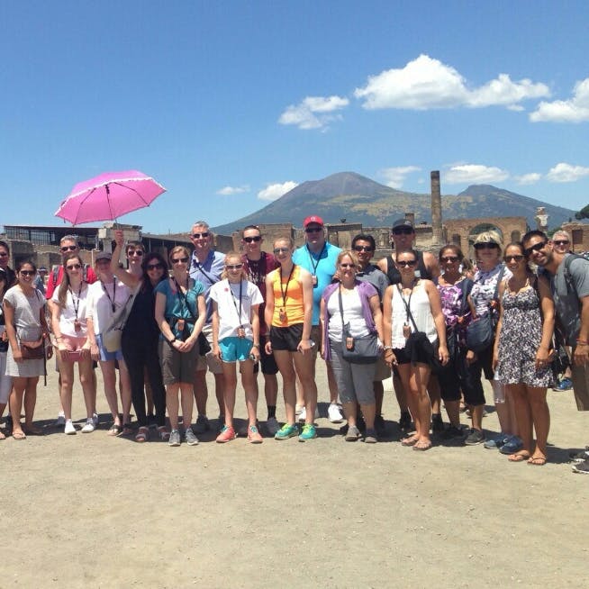 유적과 산을 배경으로 야외에 서 있는 사람들의 무리. 한 사람이 분홍색 우산을 들고 있습니다.