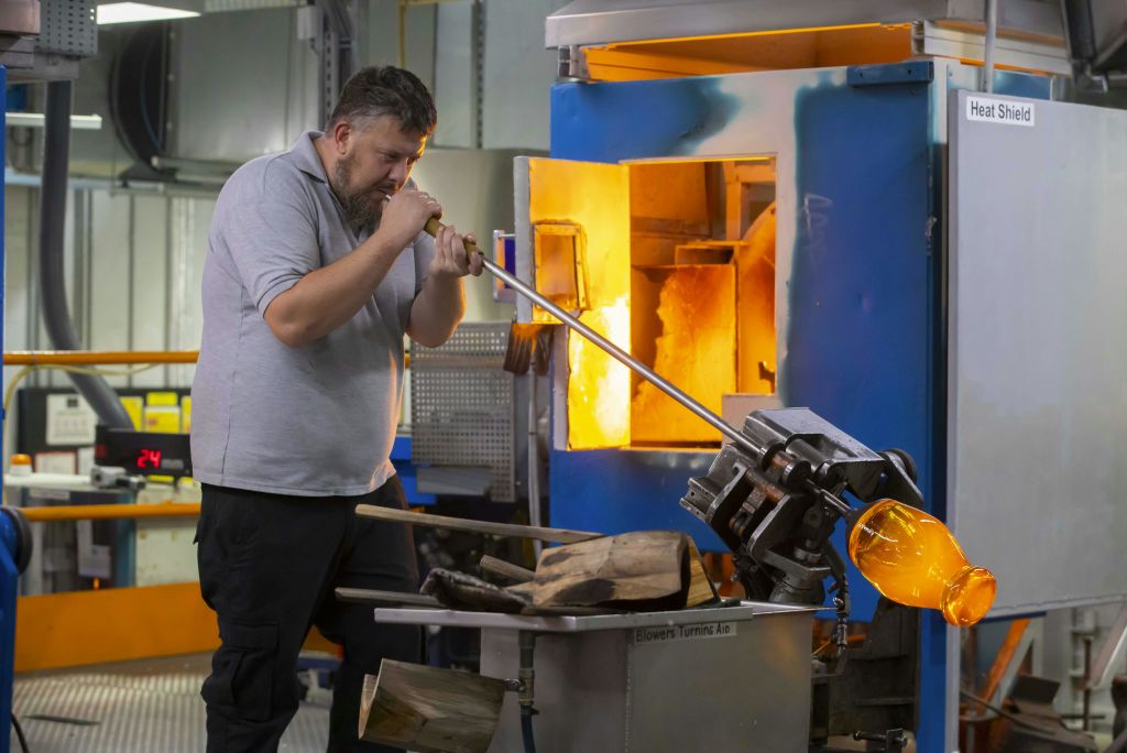 한 남성이 작업장에서 긴 파이프를 사용하여 유리를 불고 있으며, 근처에 도구와 장비가 있는 유리로 옆에 서 있습니다.
