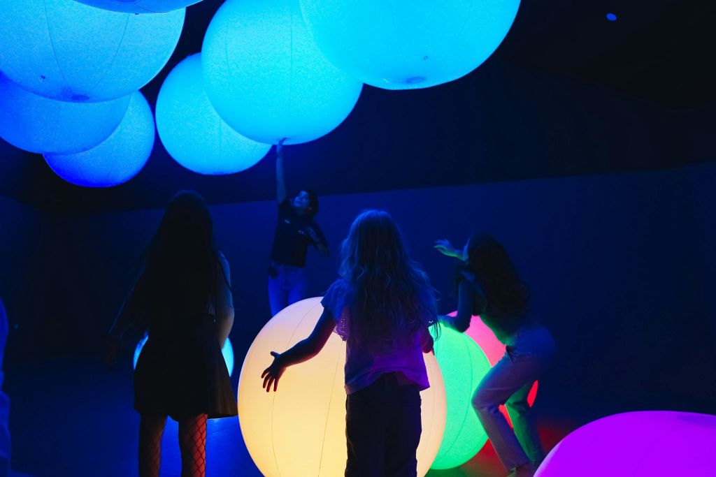 한 무리의 사람들이 파란색과 여러 가지 빛깔의 조명으로 빛나는 어두운 방에서 크고 빛나는 팽창식 구와 상호 작용합니다.
