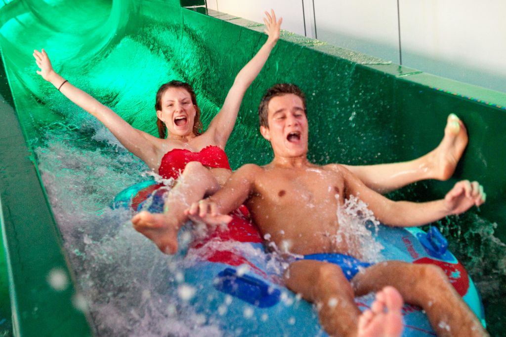 수영복을 입은 두 사람이 팽창식 뗏목에서 워터 슬라이드를 미끄러지며 팔을 들고 미소를 짓고 주위에 물이 튀고 있습니다.