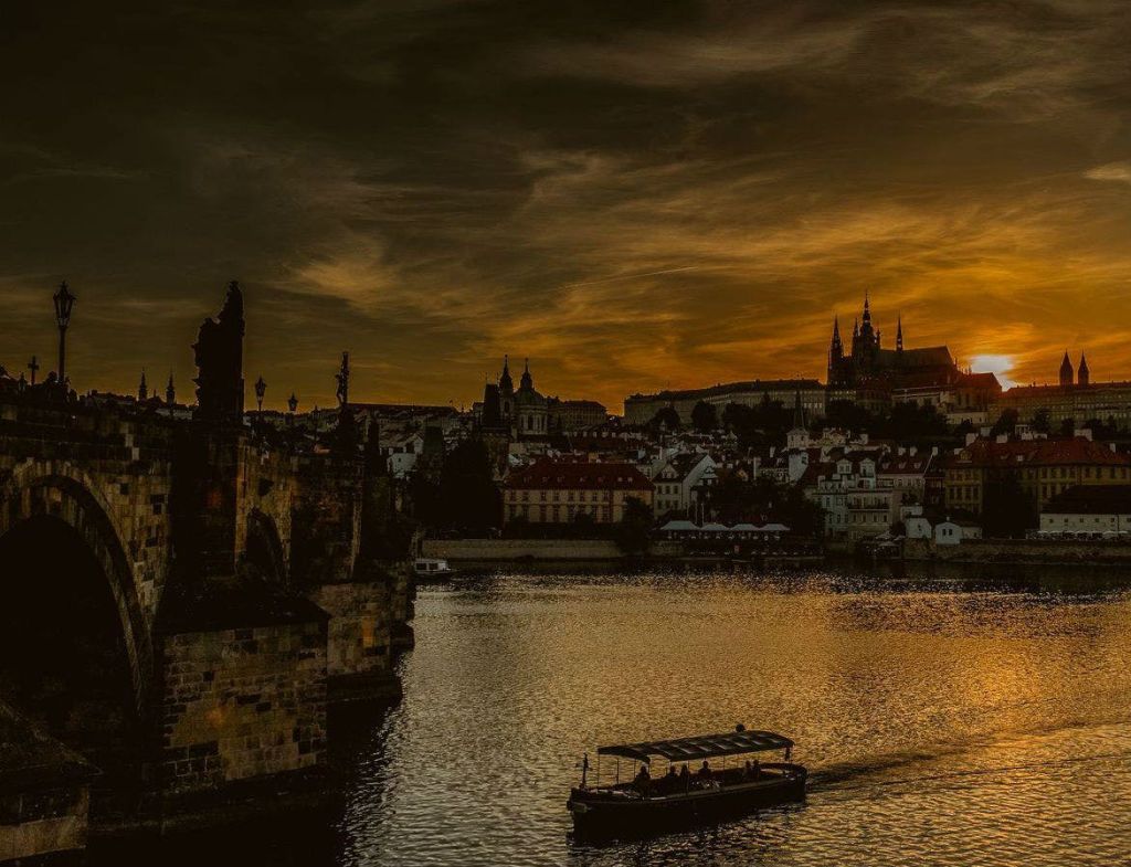 드라마틱한 하늘 아래에서 보트, 역사적인 건물, 다리가 있는 강 위로 석양을 즐겨보세요.