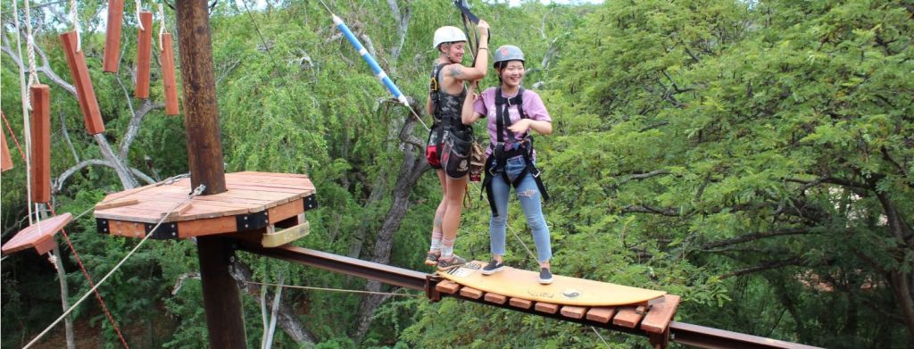 안전 장비와 헬멧을 착용한 두 사람이 울창한 푸른 숲의 나무 다리에 서 있습니다.
