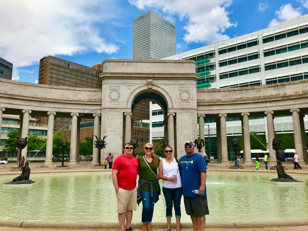 화창한 날 높은 건물이 보이는 분수와 아치 구조물 앞에 네 사람이 서 있습니다.