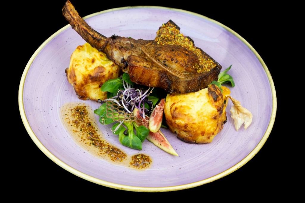 그릴에 구운 미트 찹, 구운 감자 2개, 채소, 무화과 슬라이스, 소스 드리즐이 담긴 도금 접시를 보라색 접시에 담았습니다.