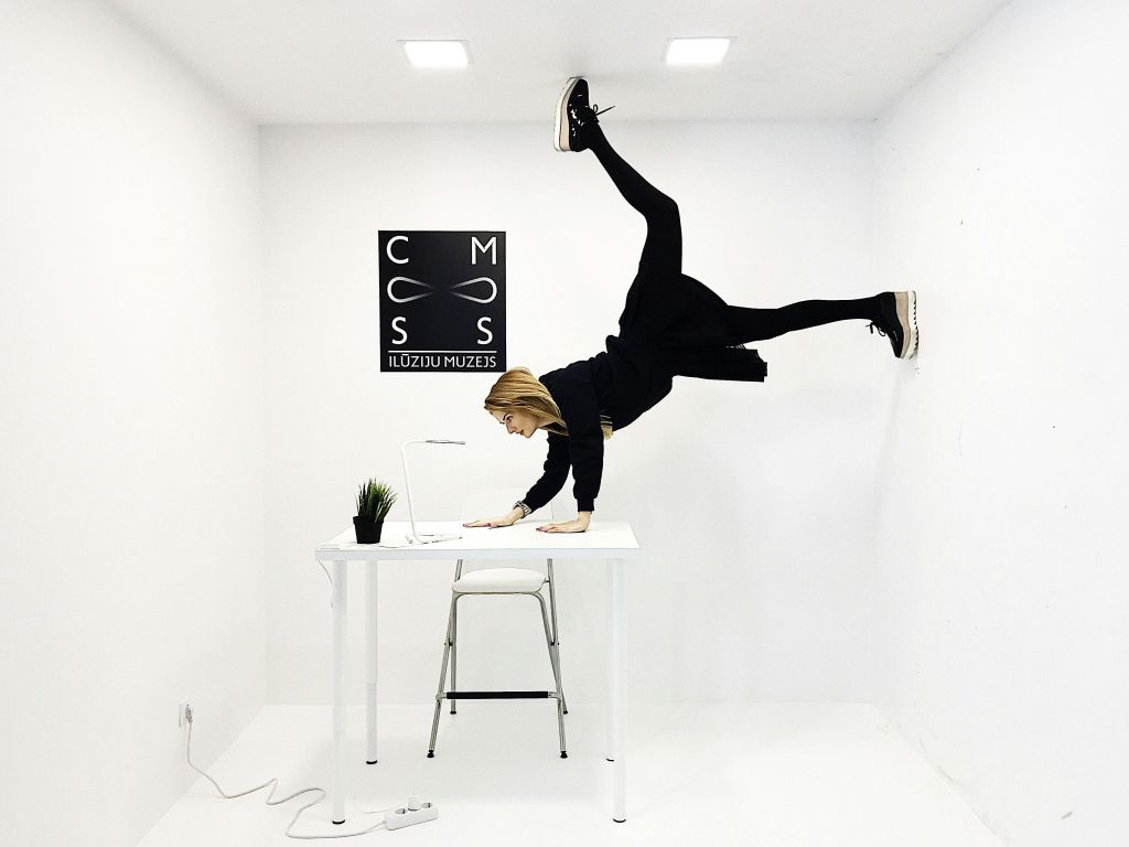 벽에 모노크롬 일루젼 박물관 간판이 있는 흰색 방의 테이블에 한 여성이 손잡이를 놓고 있습니다.