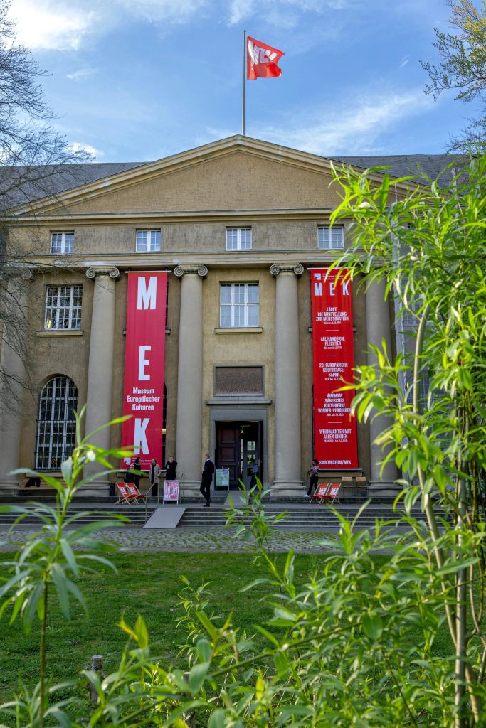 커다란 기둥과 입구에서 "MEK" 를 읽는 빨간 배너가 있는 고전적인 건물입니다. 두 사람이 들어오고 있습니다.