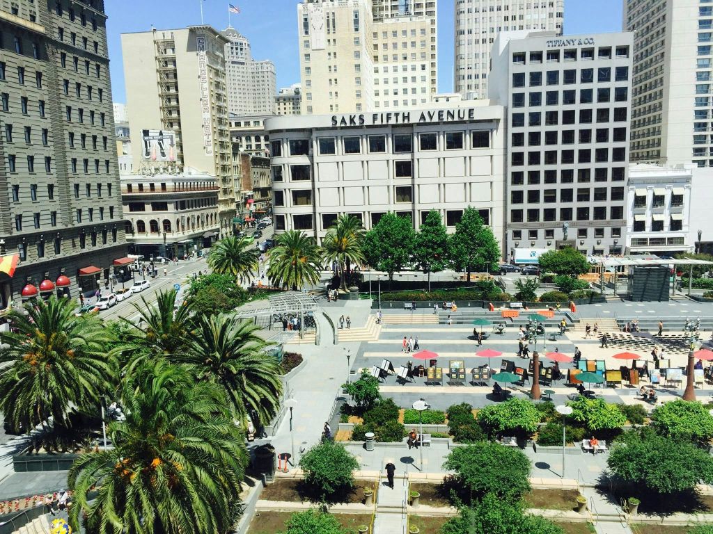 삭스 피프스 애비뉴 (Saks Fifth Avenue) 와 티파니 앤 컴퍼니 (Tiffany & Co.) 를 포함한 높은 건물로 둘러싸인 사람들, 녹지, 야외 좌석이 있는 도심 공원.