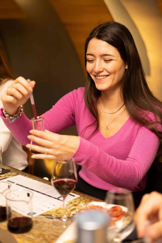 분홍색 스웨터를 입은 여성이 점적기를 사용하여 실내 유리 용기에 액체를 옮기면서 미소를 짓고 있습니다.