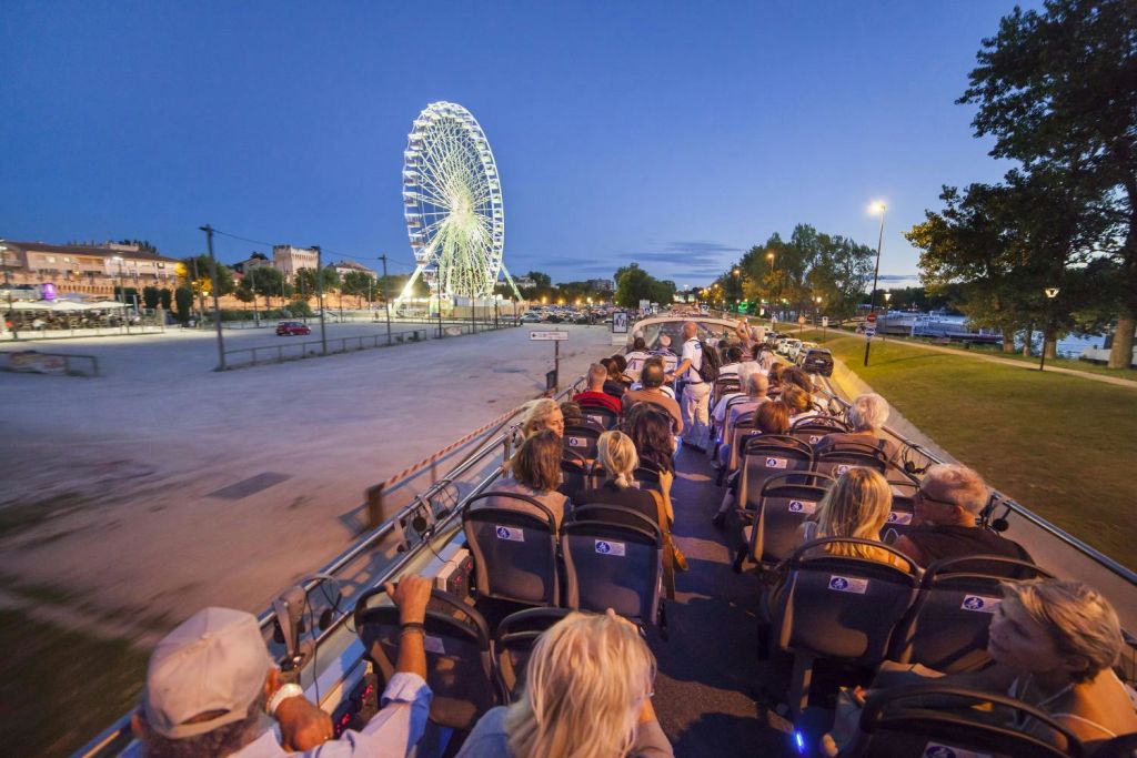 해질 무렵 오픈탑 버스에 앉아 밝은 대관람차로 향하는 사람들. 나무와 가로등도 보입니다.