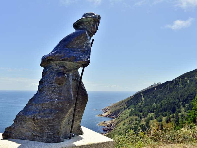 맑고 푸른 하늘 아래 나무와 절벽이 있는 해안 풍경이 내려다보이는 등산객의 청동 동상.