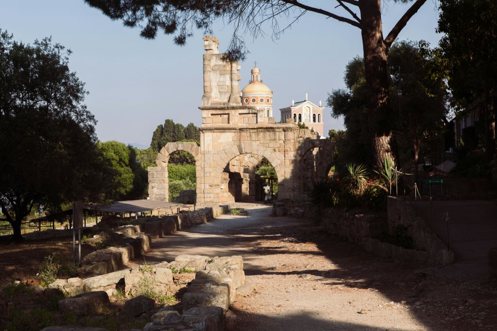 통로가 있는 고대 석조 아치길. 나무와 녹지로 둘러싸인 두 개의 돔형 건물이 배경입니다.