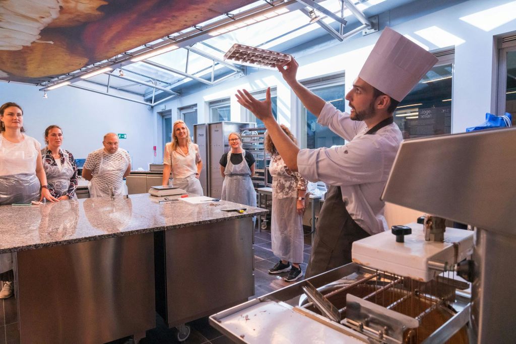 앞치마를 입은 한 무리의 사람들이 현대적인 주방에서 강사가 요리 기법을 시연하는 모습을 세심하게 지켜보고 있습니다.