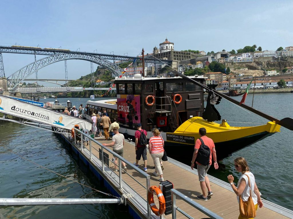 커다란 다리와 언덕 위 건물을 배경으로 화려한 외관이 돋보이는 보트를 타고 선착장을 걷는 사람들.