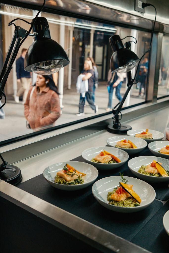 건물 내부의 두 개의 탁상용 램프 아래에 음식이 놓여 있으며, 창문으로 보행자가 보입니다.