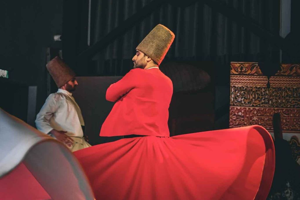 조명이 희미한 무대에서 빨간색과 흰색 가운과 키가 큰 모자를 쓰고 수피 전통 회오리를 연주하는 두 사람.