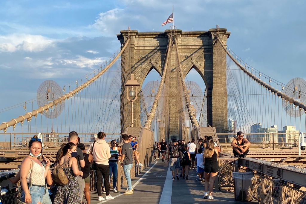 미국 국기를 배경으로 푸른 하늘 아래 브루클린 다리를 걸으며 사진을 찍는 사람들.