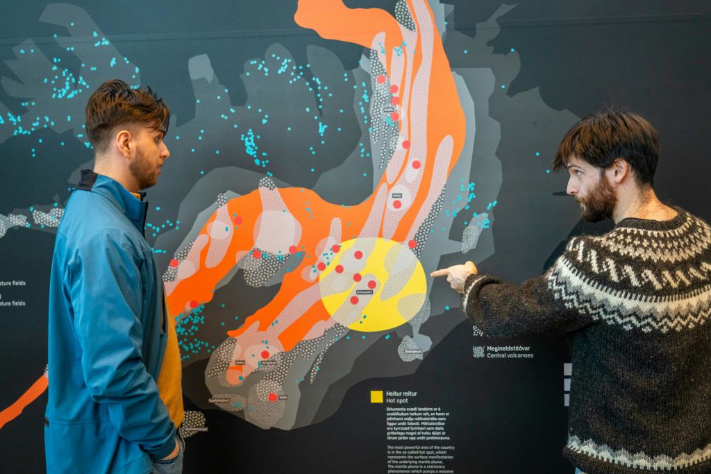 파란색 재킷을 입은 한 남성이 벽에 걸린 다채로운 지형도를 바라보고 있습니다. 다른 사람의 손이 지도를 가리킵니다.