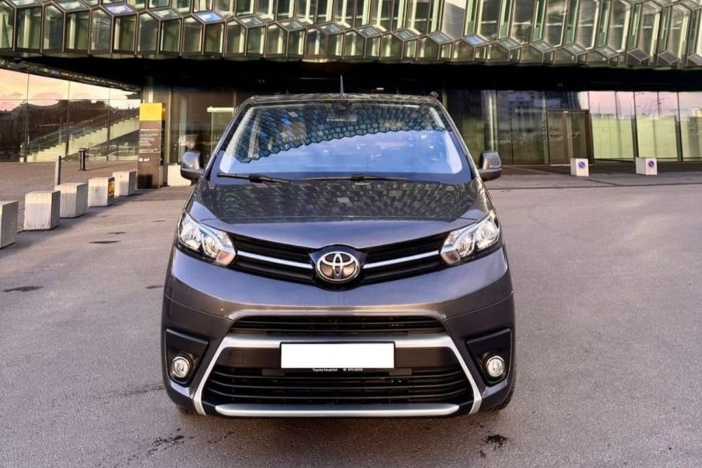 현대적인 유리 건물을 배경으로 거리에 주차된 회색 도요타 미니밴의 전면 모습.