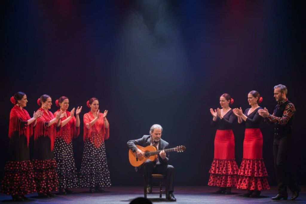 기타리스트가 무대 위에 앉아 빨간색과 검은색 의상을 입은 플라멩코 댄서들에게 둘러싸여 리듬에 맞춰 박수를 치고 있습니다.