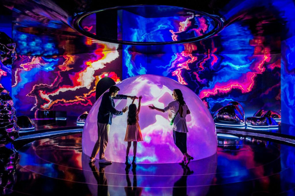 벽에 선명하고 화려한 추상적 조명이 투영된 방에서 세 사람이 조명 돔과 상호 작용합니다.