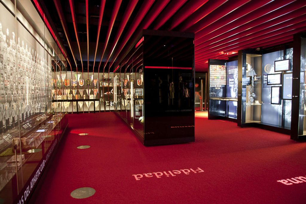 레드 카펫, 블랙 디스플레이 케이스 및 다양한 유물이 전시된 박물관 전시관입니다. 천장에는 빨간색 조명과 평행 빔이 있습니다.