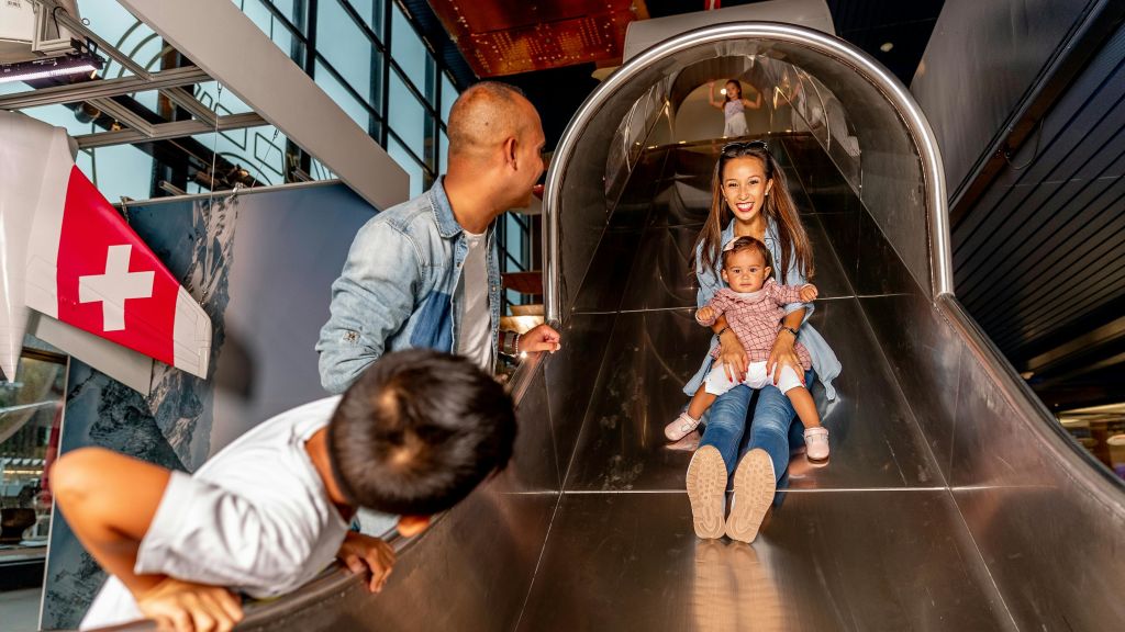 한 여성과 한 유아가 금속으로 된 놀이터 슬라이드를 미끄러지듯 내려가고 있습니다. 한 남자가 옆으로 서서 그들을 바라보며 미소 짓고 있습니다.