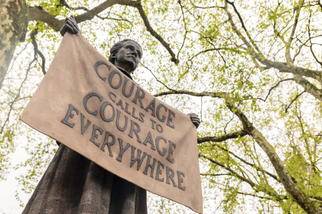 나뭇가지와 나뭇잎을 배경으로 "용기는 어디서나 용기를 부른다" 라는 표지판을 들고 있는 동상.