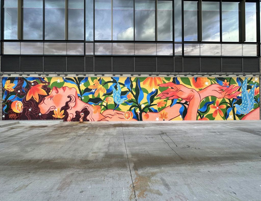 커다란 창문이 있는 현대적인 건물과 맞닿아 있는 꽃과 과일로 둘러싸인 안락한 모습의 다채로운 벽화입니다.