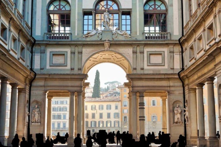 실루엣의 사람들이 기둥과 조각이 있는 웅장한 건물의 아치형 통로를 지나 도시 건물의 햇살이 내리쬐는 전망으로 이어집니다.