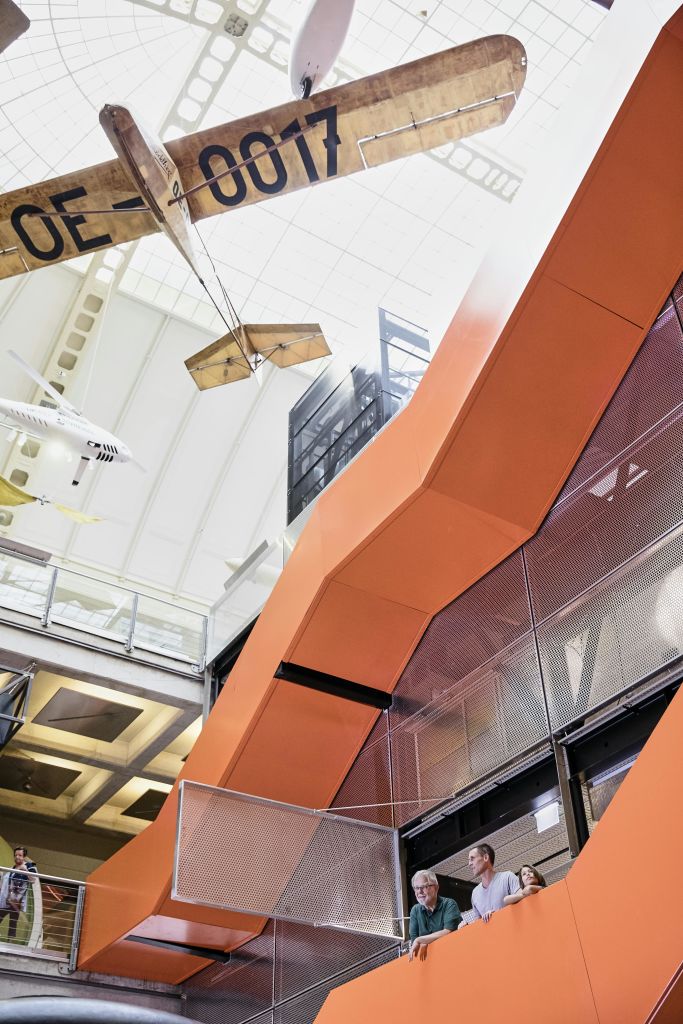 주황색 구조, 매달린 비행기 모형, 채광창이 있는 높은 천장이 있는 현대적인 건물의 내부.