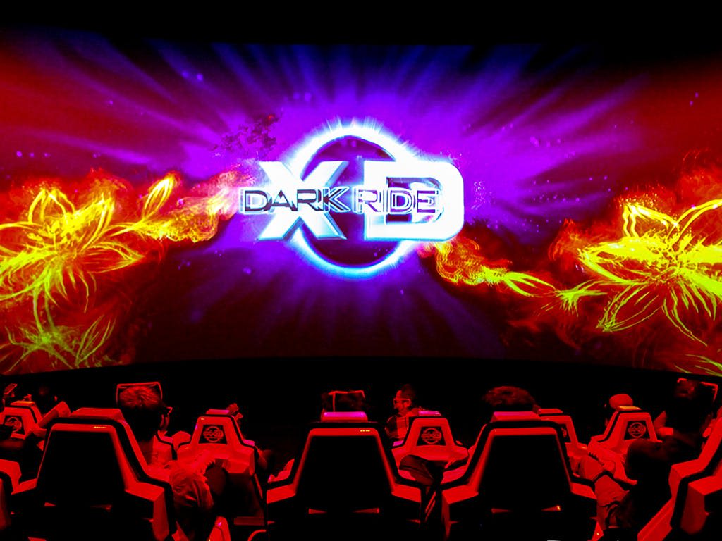 모션 시뮬레이터 의자에 앉아 "XD Dark Ride" 라는 제목의 영화 화면을 불타는 그래픽으로 감상하는 사람들.
