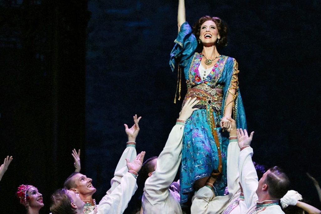 화려한 색상의 드레스를 입은 여성이 희미하게 빛나는 무대에서 연극 공연 중 여러 공연자가 들어 올립니다.