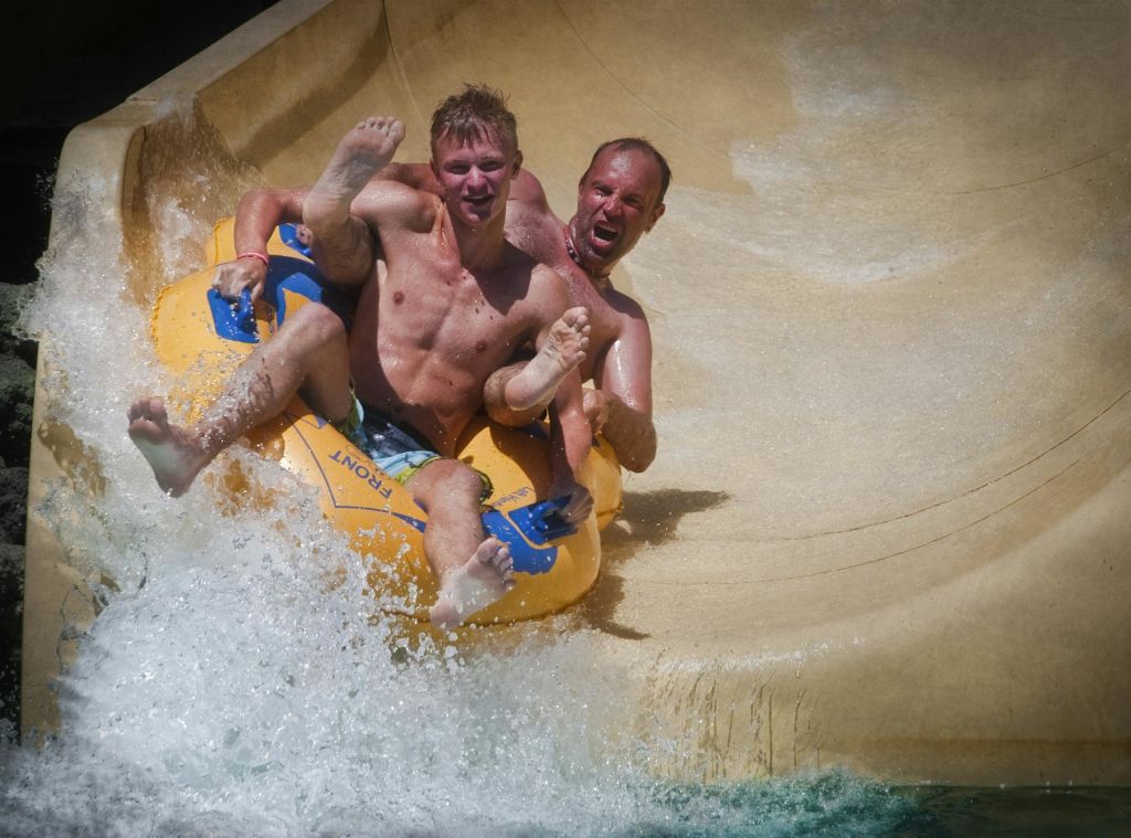 노란색 팽창식 뗏목을 탄 두 남성이 워터 슬라이드를 따라 미끄러져 내려오고 주위에 물이 튀고 있습니다.