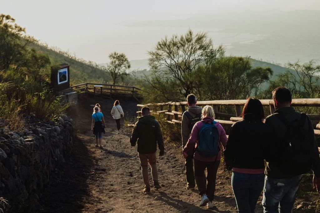 한 무리의 사람들이 나무 울타리와 드문드문 나무를 경계로 언덕길을 따라 흙길을 걷고 있습니다.
