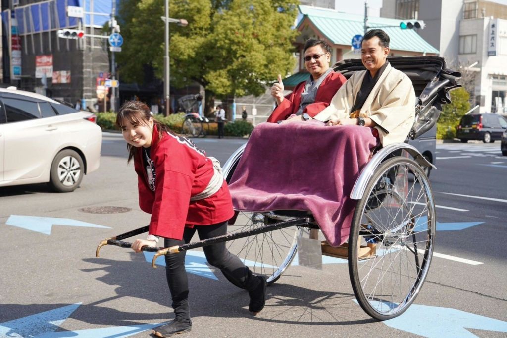 햇살이 내리쬐는 거리에서 한 여성이 전통 의상을 입은 웃는 두 남성을 태운 인력거를 끌고 있습니다.