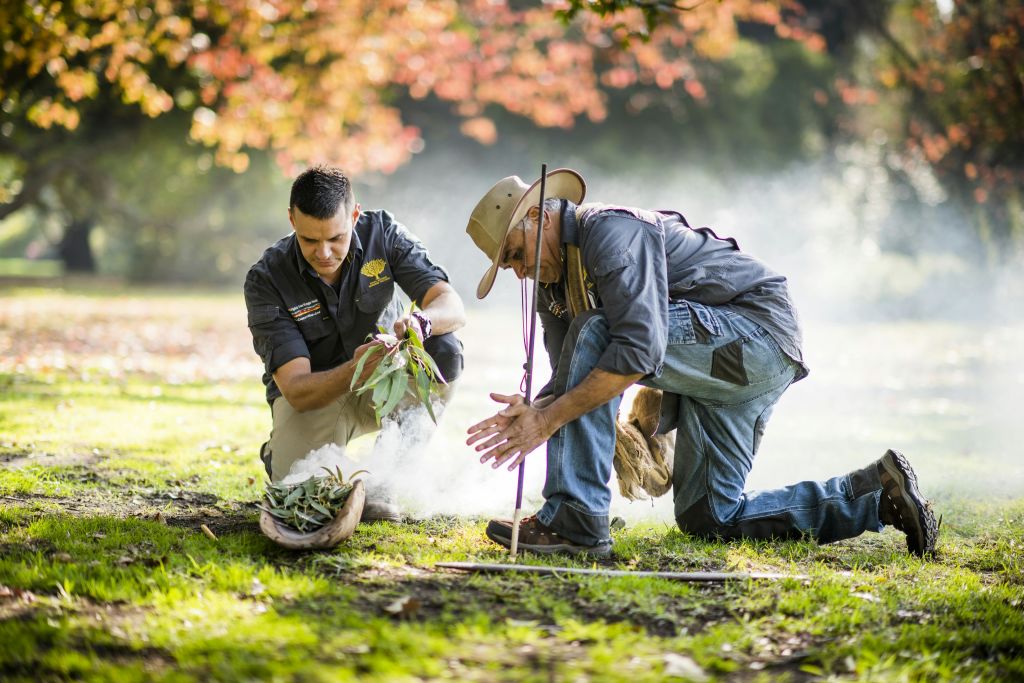 두 남자가 풀밭에 무릎을 꿇고 나뭇잎으로 흡연 의식을 합니다. 붉은 잎을 배경으로 한 가을 나무.